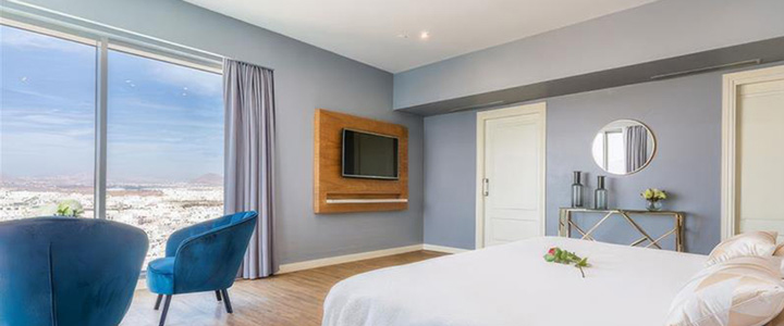 Elegantes Schlafzimmer in Blautönen mit einem großen Doppelbett, zwei samtenen Sesseln am Fenster und einer beeindruckenden Aussicht über eine Stadtlandschaft bis hin zu Bergen.