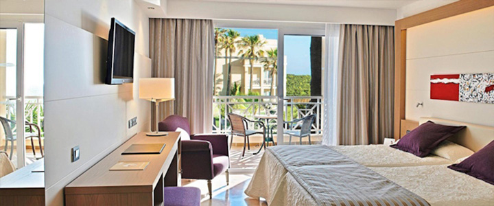 Das Doppelzimmer im TUI BLUE Playa La Barrosa ist hell und modern eingerichtet. Durch die Balkontür fällt der Blick auf Palmen und den grünen Außenbereich.